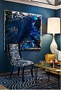 paint colors for living room, dark blue walls, blue and cream carpet, lighter blue and cream chair with black details, white lamp and glass table with gold details, large dark blue abstract painting